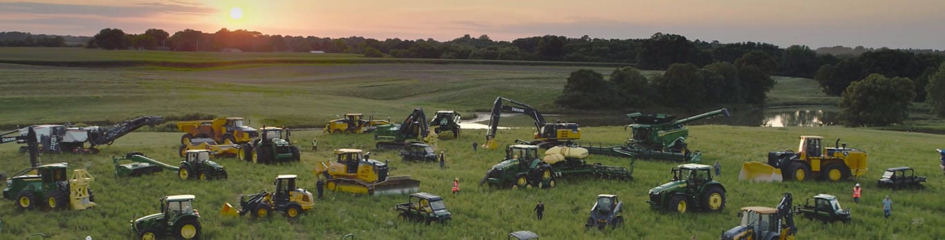 Persone che camminano tra macchine John Deere di vario tipo su un grande campo.