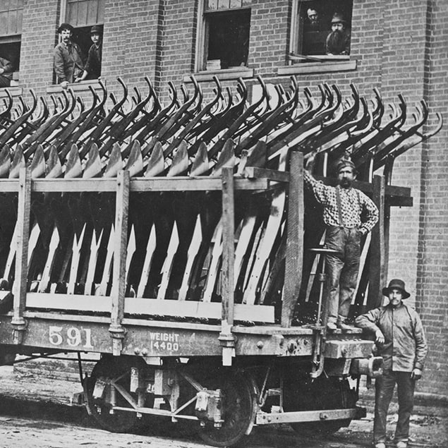 Fotografia del 1882 raffigurante tre uomini in piedi accanto al carrello ferroviario Deere & Co carico di aratri in acciaio pronti per essere spediti e operai che guardano fuori dalla finestra dell'edificio retrostante