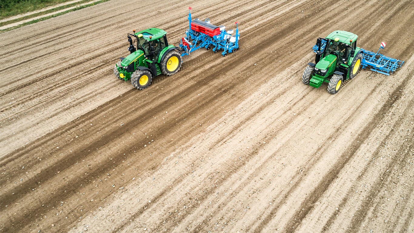 Due trattori John Deere 6M su un campo di terra spoglia. Uno traina una seminatrice Monsomen. L'altro traina una combinazione di letti di semina Lemken