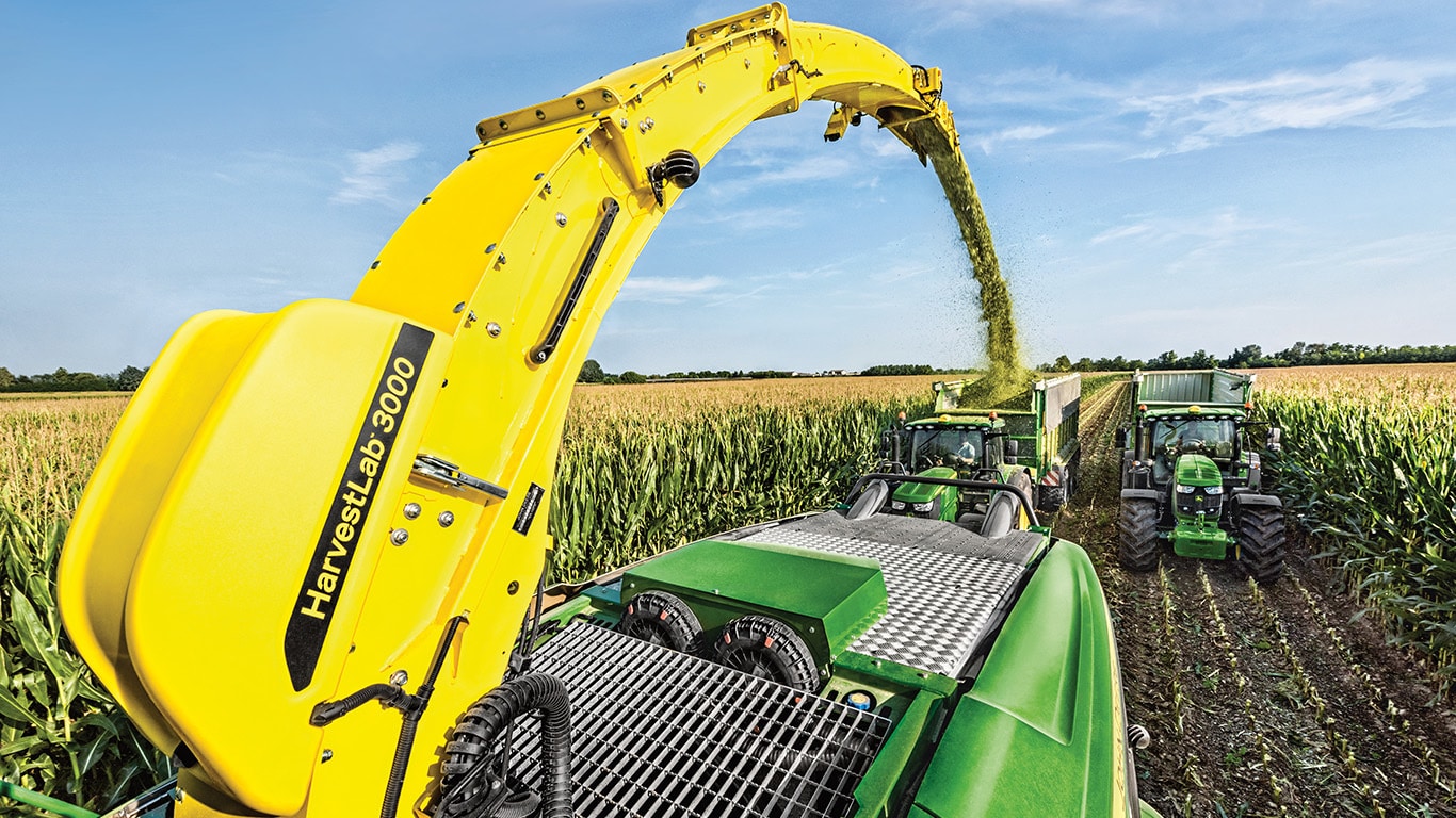 Vista dal retro della trincia semovente John Deere Serie 9000 con due carri per insilato trainati da trattori John Deere direttamente dietro