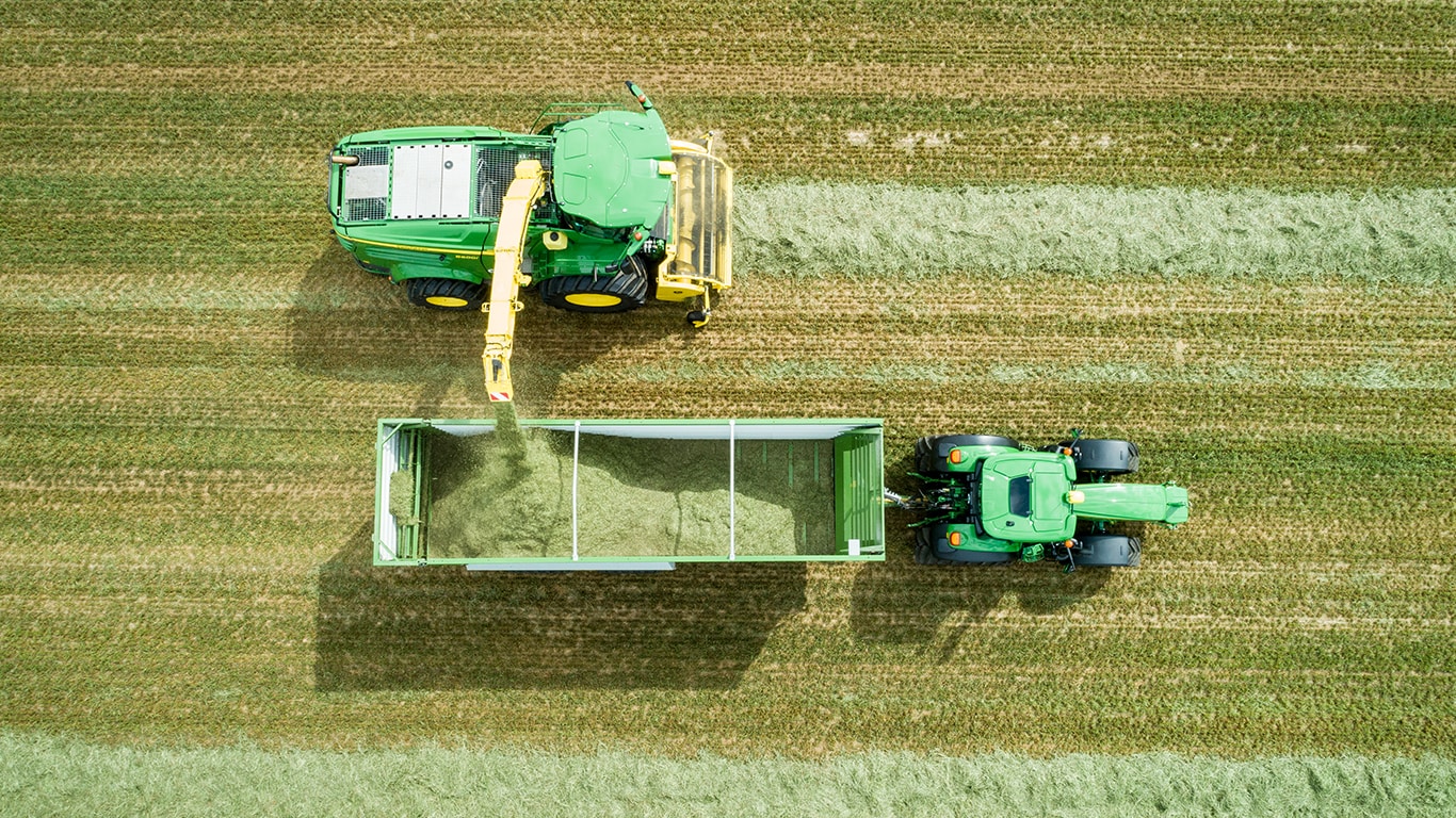 Foto aerea della trincia semovente John Deere Serie 8000 con un pick-up per l'erba che carica insilato in un carro trainato da un trattore John Deere